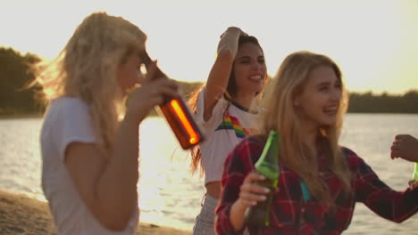 Girls-carefree-and-cheerful-move-young-bodies-on-the-beach-with-beer-on-the-open-air-party-near-the-sand-river-coast-in-summer-evening.